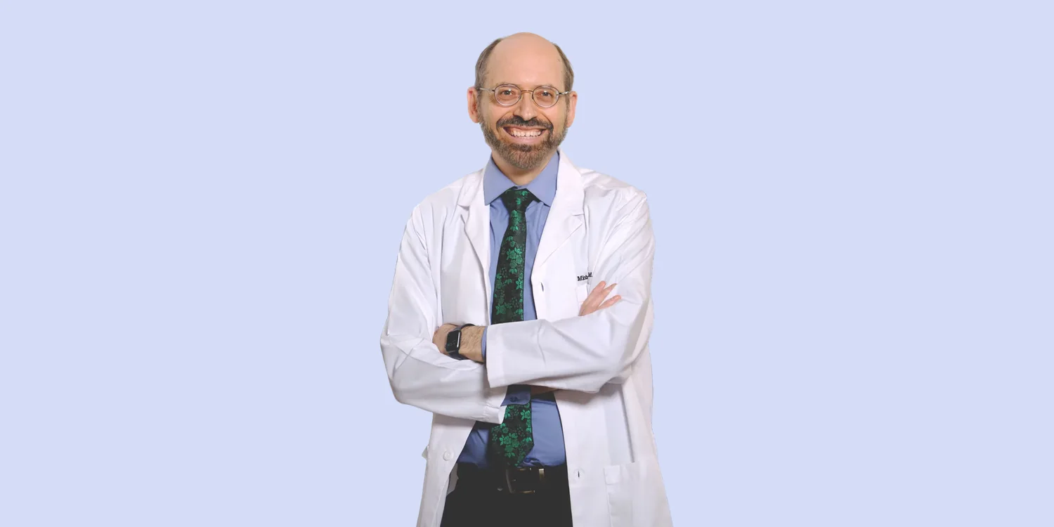 Dr Michael Greger in a white coat, arms folded and smiling, on a pale violet background