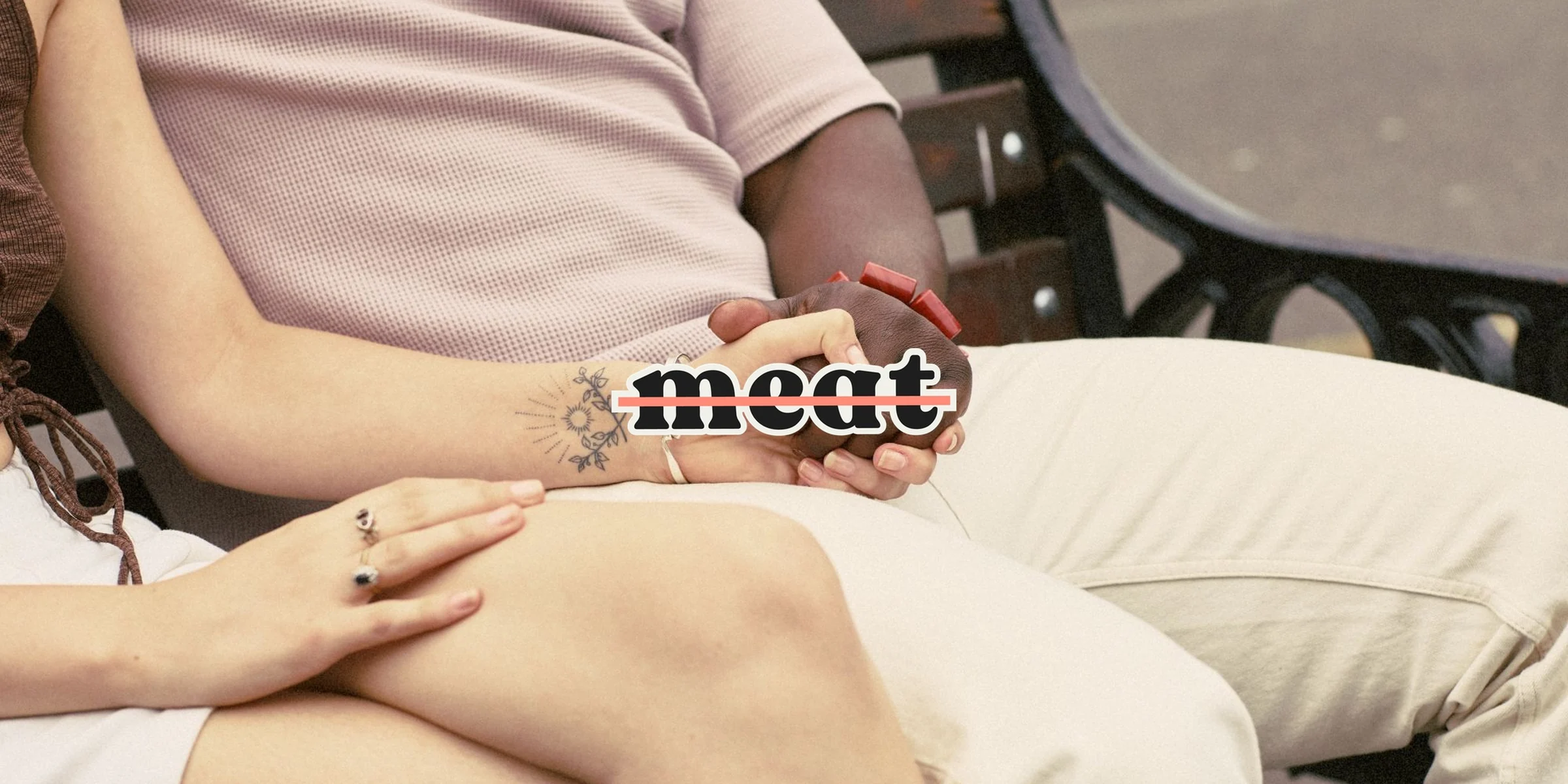 Two people holding hands on a bench, with a crossed-out "meat" sticker over their hands
