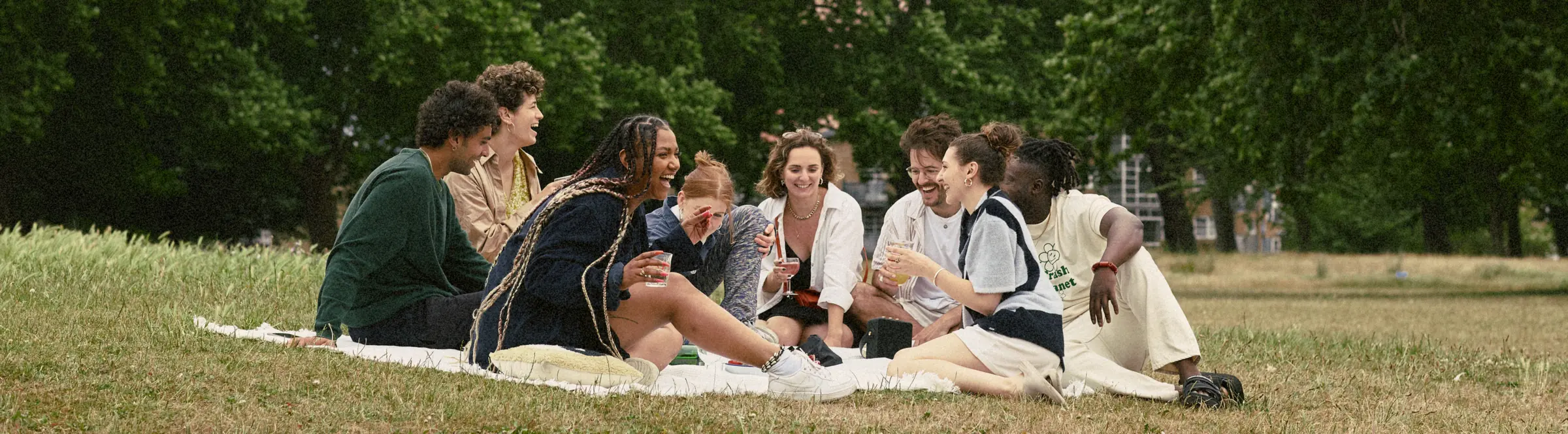 Friends laughing together on a picnic blanket in a park, with tall trees and a tower block behind them
